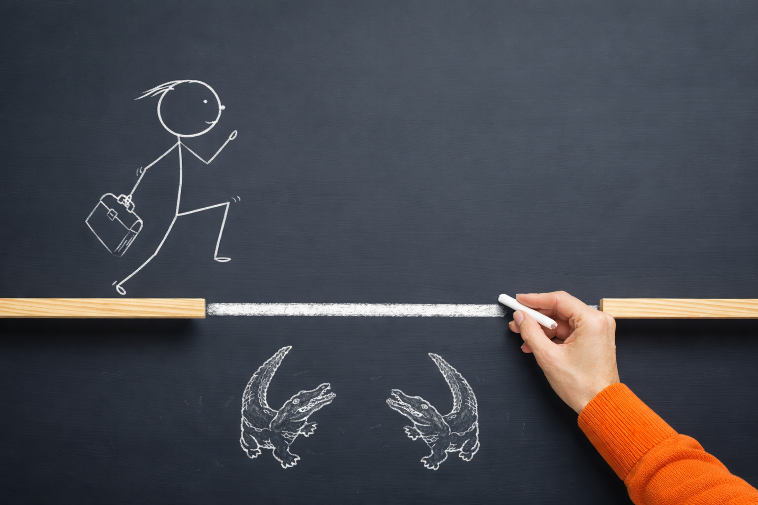 Concept image of overcoming a dangerous gap: a hand draws a chalk bridge between two platforms as a business traveler crosses above crocodiles