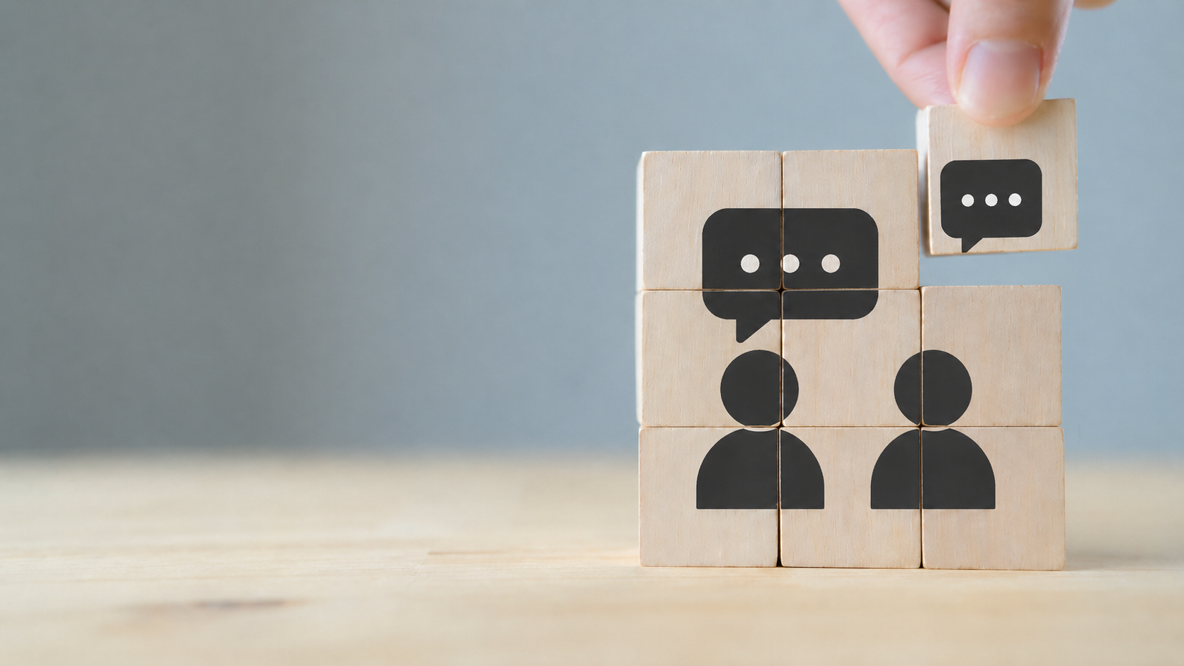 Hand placing a wooden block with a speech bubble icon above two person icons, symbolizing communication, dialogue, and customer interaction.