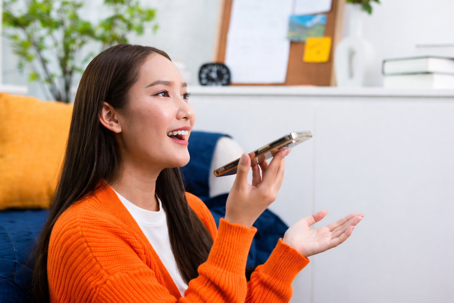 Young woman indoors smiling while speaking into AI on her smartphone, wearing an orange sweater and sitting in a bright home setting.